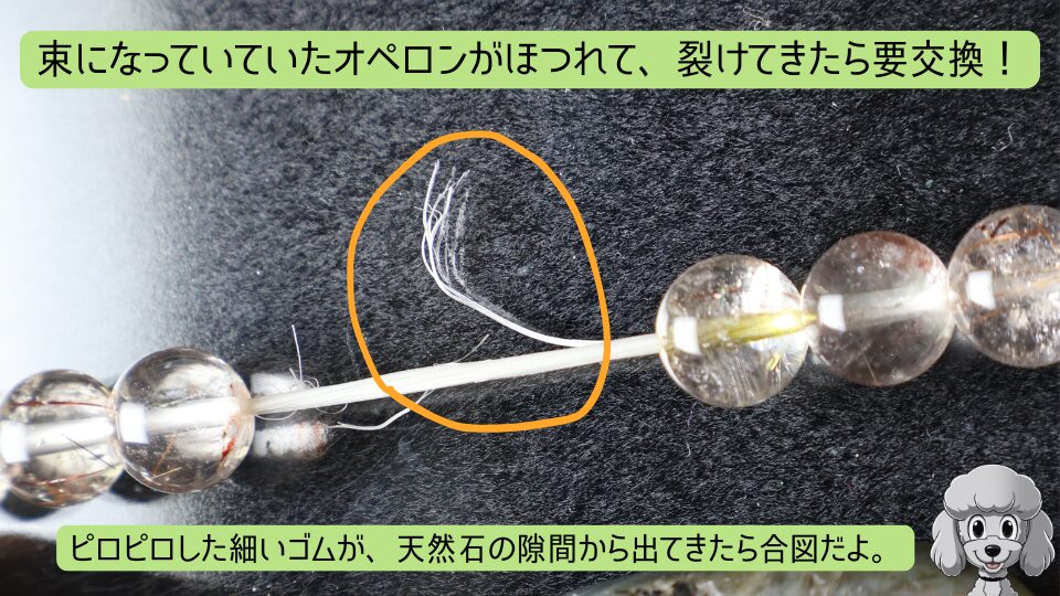 束になっていたオペロンがほつれて、裂けてきたら要交換！ピロピロした細いゴムが、天然石の隙間から出てきたら合図だよ。と書かれた、天然石ブレスレットの白いオペロンゴムがほつれている様子をオレンジ色の丸で囲んで解説する画像。右下にマスコットのおーちゃん(トイプードル)のイラスト
