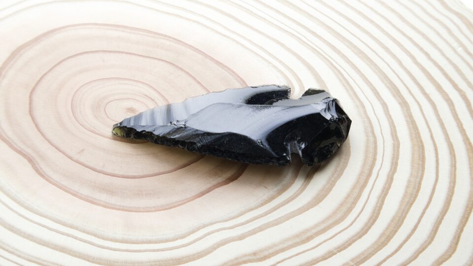 年輪がはっきりと見える丸い木のプレートの中央に置かれた、艶のある黒色をした鏃(やじり)型のオブシディアン(黒曜石)。代表だっち。が妻・えむまる。と結婚するきっかけにもなった、人生を180度変えた重要な天然石