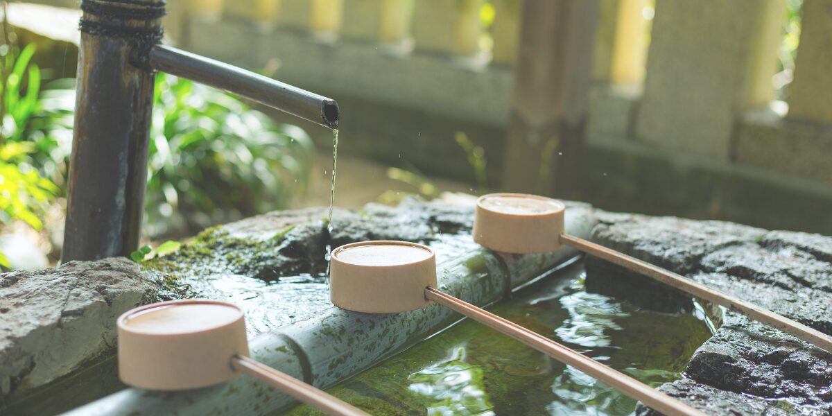 神社にある手水舎(てみずや)の風景。竹の筒から水が流れ落ち、木製の柄杓(ひしゃく)が置かれた石の鉢に緑の木漏れ日が差し込んでいる様子